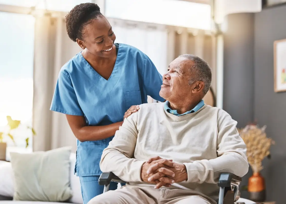A healthcare worker in blue scrubs smiles and places a hand on the shoulder of a seated older man. The man looks up at her, smiling, in a well-lit room with plants in the background.